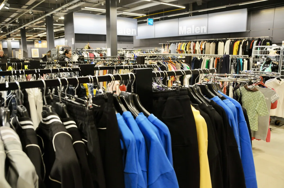 Auf der linken Seite sind neue, teils exklusive Markenkleider, auf der rechten Seite gebrauchte Kleider. Man sieht Kleider an Stangen hängen.